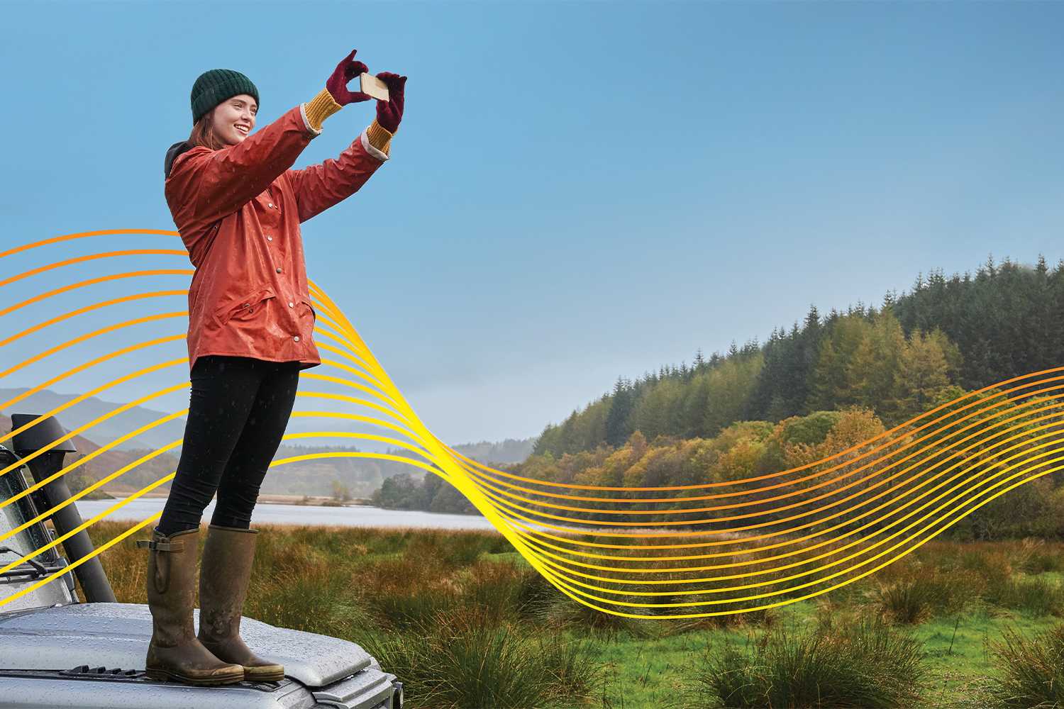 Woman standing on the hood of an old car, in a rural location with mountains in background, holding up her phone to take a photo, with "wave" graphic imposed over the image