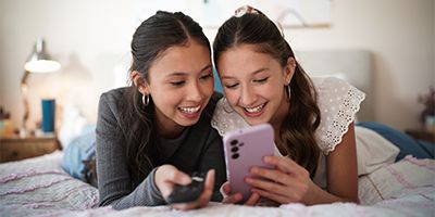 Two females lying on a bed looking at a smartphone together, smiling